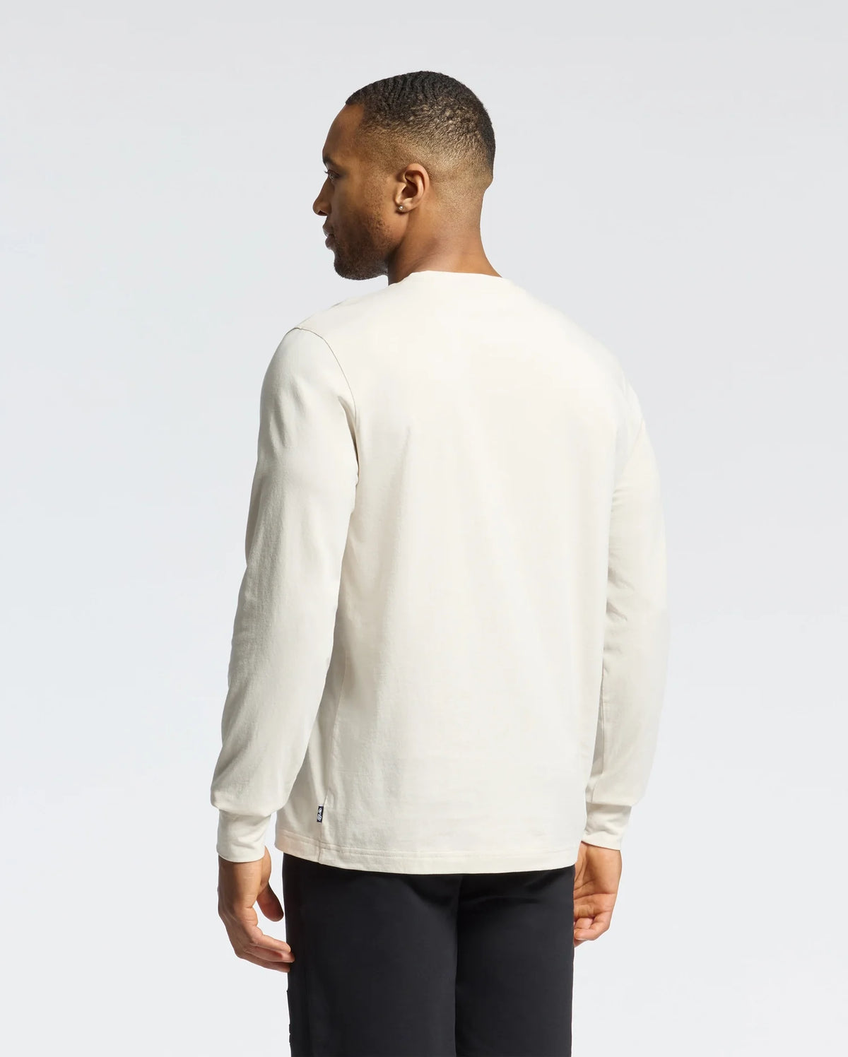 A person in a Psycho Bunny MENS ALBERTON LONG SLEEVE TEE (B6T867D200), white Pima cotton, and black pants stands against a plain background, facing away from the camera, looking to the right.