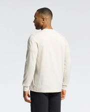 A person in a Psycho Bunny MENS ALBERTON LONG SLEEVE TEE (B6T867D200), white Pima cotton, and black pants stands against a plain background, facing away from the camera, looking to the right.