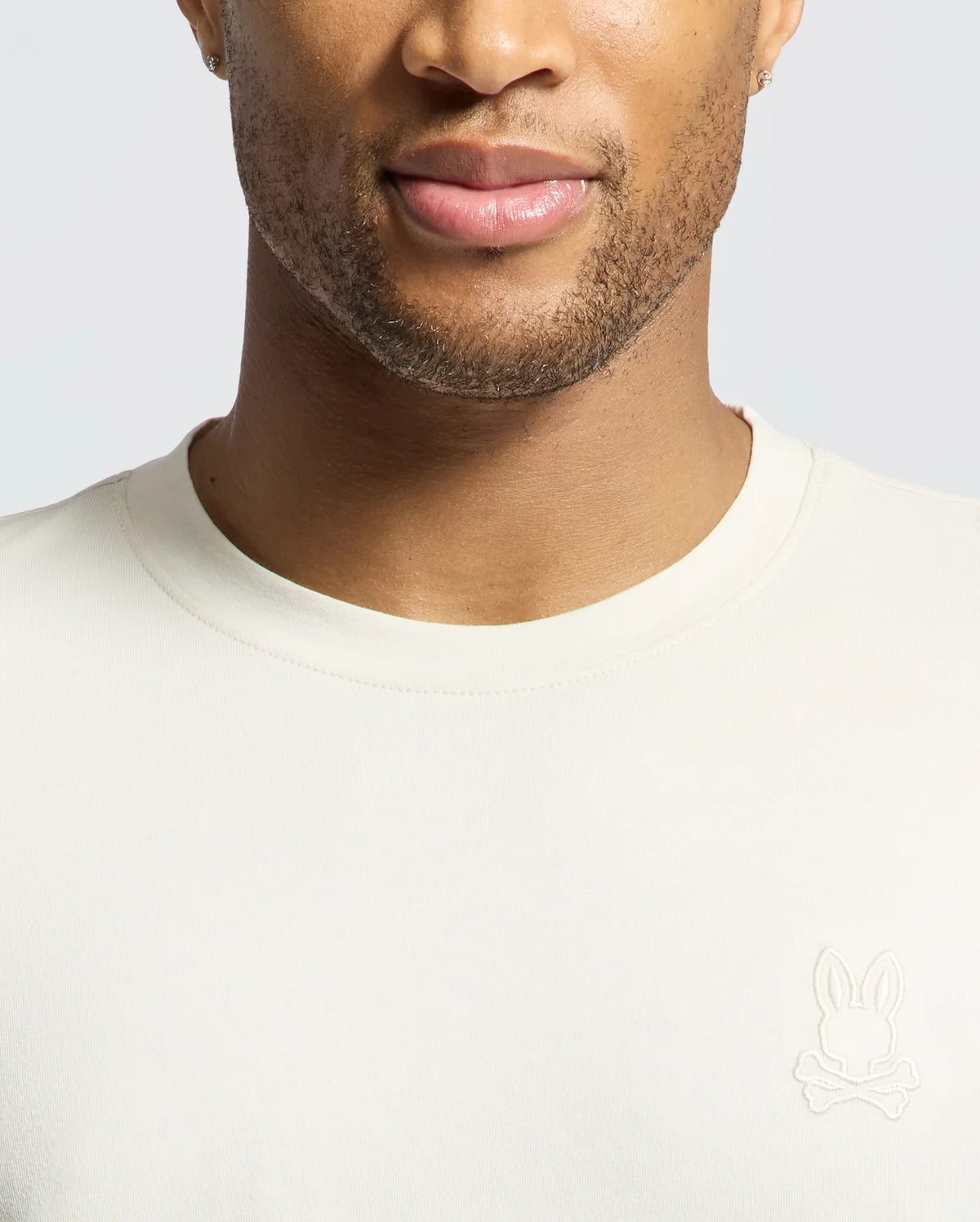 A person wearing a Psycho Bunny MENS ALBERTON LONG SLEEVE TEE in cream, with a bunny and crossbones logo on the left. Made from soft Pima cotton, it highlights the face and upper body against a light gray background, making it an ideal layering piece.