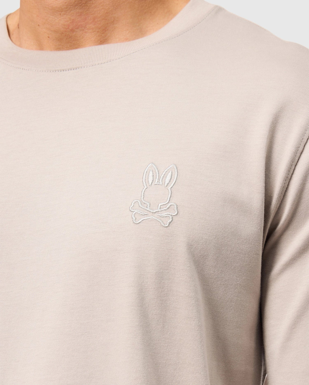 Close-up of a person wearing a Psycho Bunny Alberton long sleeve tee in beige, crafted from soft Pima cotton, featuring an embroidered bunny logo above crossed bones on the chest.