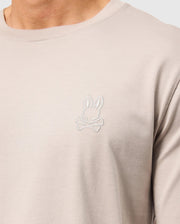 Close-up of a person wearing a Psycho Bunny Alberton long sleeve tee in beige, crafted from soft Pima cotton, featuring an embroidered bunny logo above crossed bones on the chest.