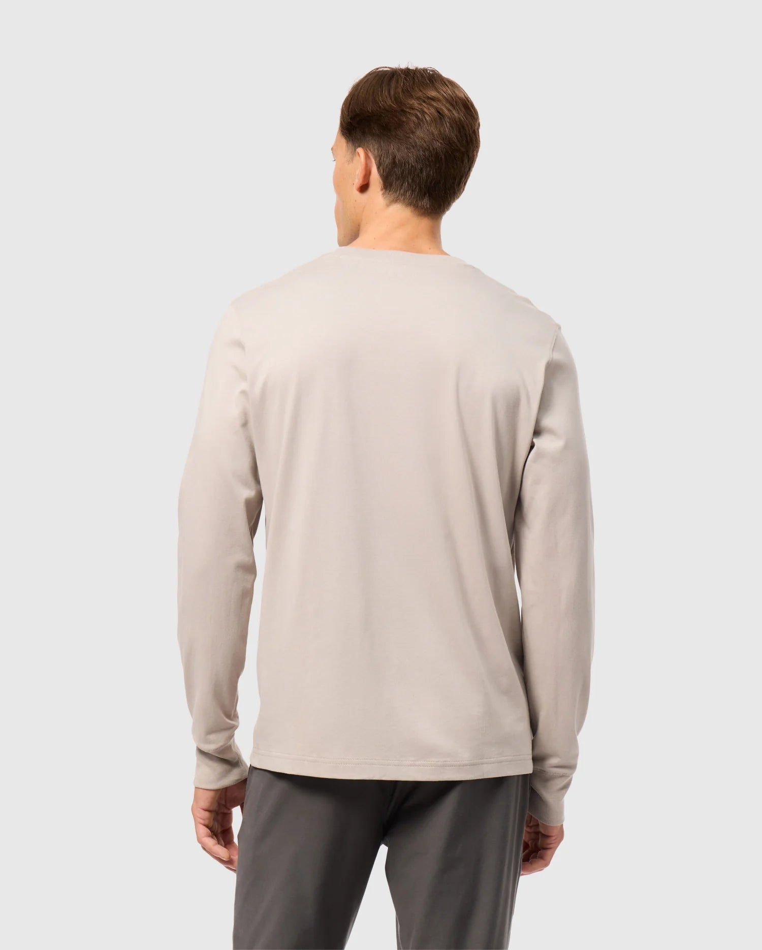 A person wearing a light beige MENS ALBERTON LONG SLEEVE TEE - B6T867D200 by Psycho Bunny and dark gray pants is standing with their back to the camera against a plain background.