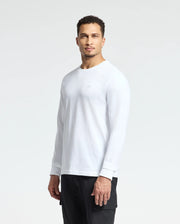A man with short curly hair is standing against a plain background, wearing the Psycho Bunny MENS ALBERTON LONG SLEEVE TEE in white, featuring a small logo on the chest, paired with black pants. His expression is neutral as he looks directly at the camera.