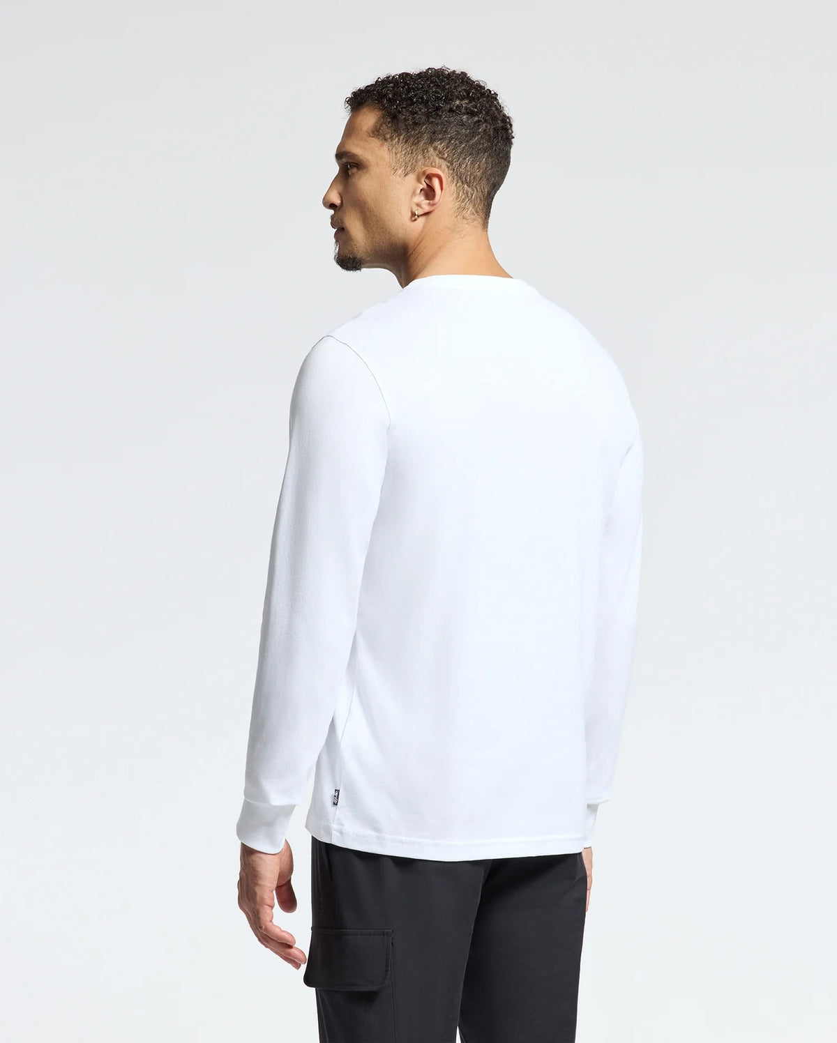 A person with short curly hair is standing slightly turned to the side, wearing a casual Psycho Bunny MENS ALBERTON LONG SLEEVE TEE - B6T867E200 and black pants. The pima cotton shirt adds a touch of elegance against the plain white background.