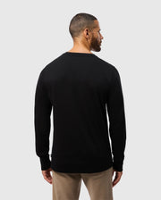 A man with short hair and a beard stands with his back to the camera, wearing the Psycho Bunny MENS MONROE LONG SLEEVE TEE in black and regular fit beige pants. The background is plain and light gray.