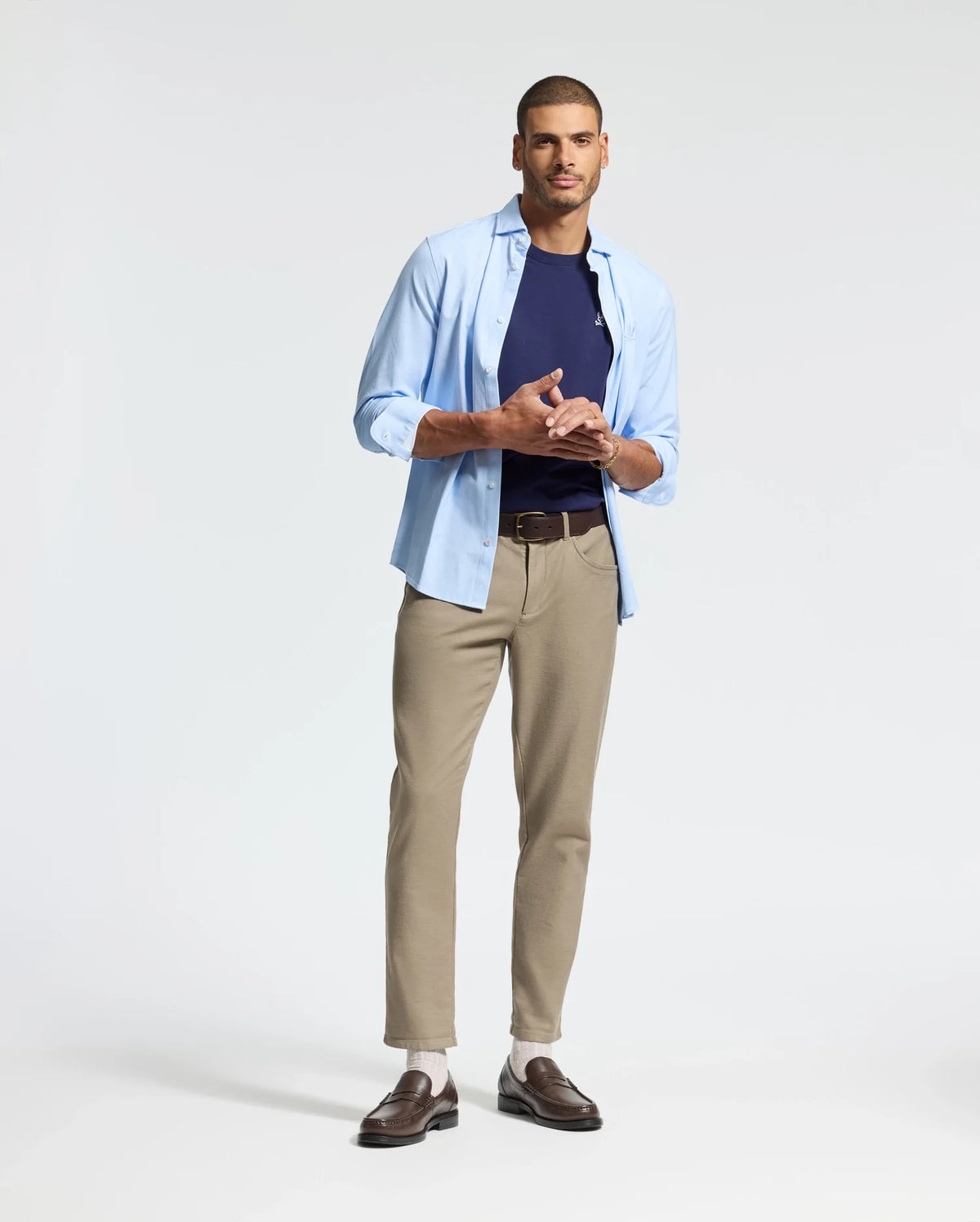 A man stands against a plain background wearing an open light blue shirt over the Psycho Bunny MENS CLASSIC CREW NECK TEE - B6U014CRPC, complemented by beige pants and brown shoes. Casually holding his hands together, he embodies a perfect blend of style and ease.