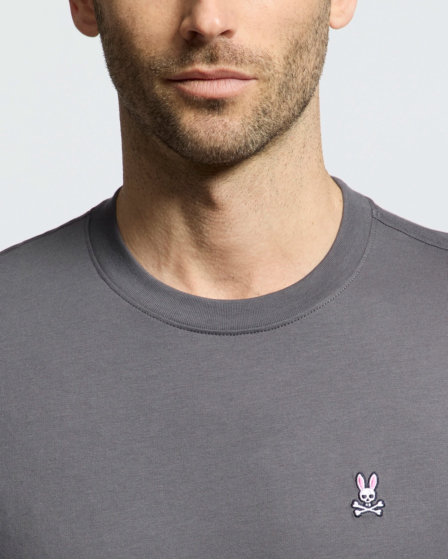 A man wears a Psycho Bunny MENS CLASSIC CREW NECK TEE (B6U014F200), a versatile gray pima cotton tee with a small pink bunny crossed-bones logo at the bottom right, captured shoulders up against a plain, light backdrop.
