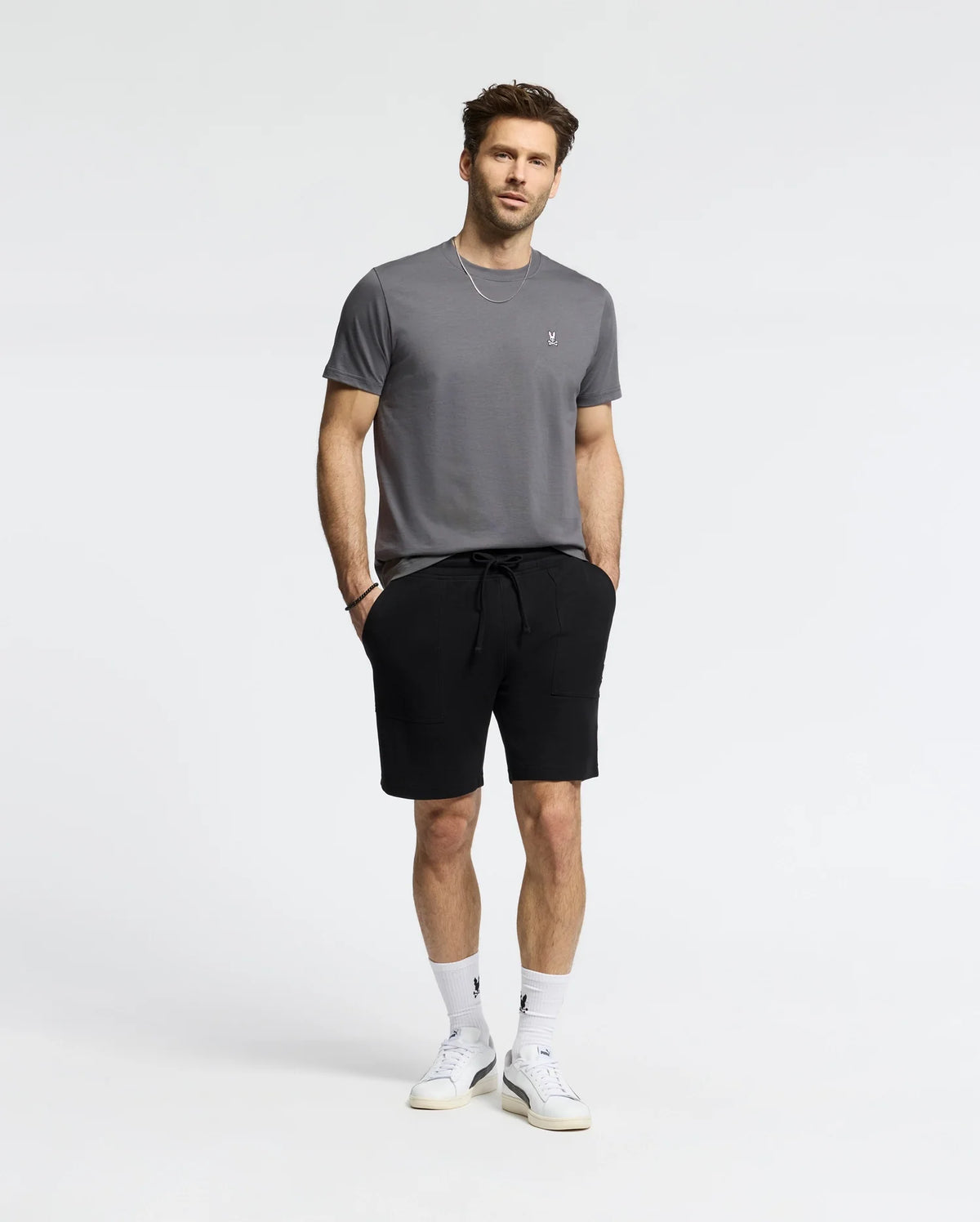 A man stands against a plain background in a Psycho Bunny MENS CLASSIC CREW NECK TEE in gray, black shorts, white socks, and white sneakers. His short, styled hair complements the relaxed look as he casually tucks his hands into his pockets.