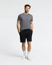 A man stands against a plain background in a Psycho Bunny MENS CLASSIC CREW NECK TEE in gray, black shorts, white socks, and white sneakers. His short, styled hair complements the relaxed look as he casually tucks his hands into his pockets.