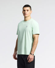A person with short hair stands slightly to the side wearing Psycho Bunny's MENS CLASSIC CREW NECK TEE - B6U014F200 in light green, featuring an embroidered Bunny logo on the chest. They pair it with black pants and a watch against a plain grayish-white background.
