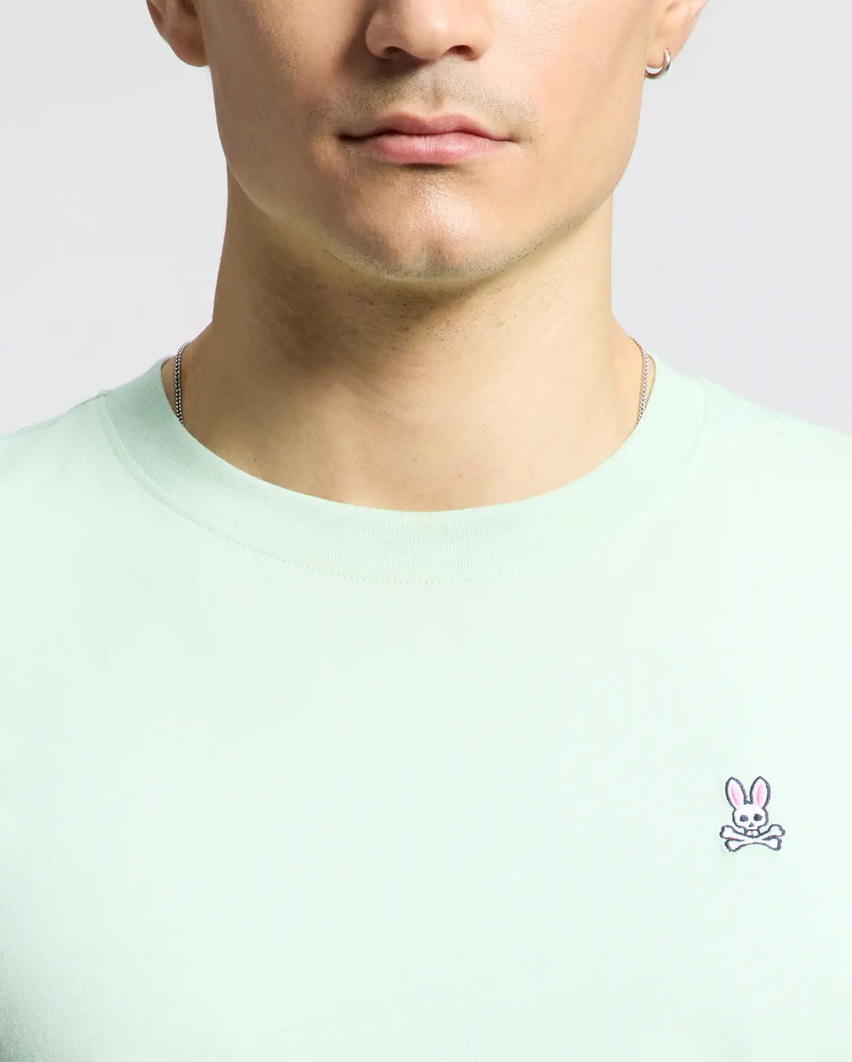 A person in a Psycho Bunny light green MENS CLASSIC CREW NECK TEE - B6U014F200, made of soft Pima cotton with an embroidered Bunny logo with pink ears, is depicted. They have short hair, a necklace, and an earring; the image is cropped to show only the lower face and upper torso.