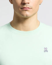 A person in a Psycho Bunny light green MENS CLASSIC CREW NECK TEE - B6U014F200, made of soft Pima cotton with an embroidered Bunny logo with pink ears, is depicted. They have short hair, a necklace, and an earring; the image is cropped to show only the lower face and upper torso.