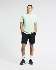 A person stands against a plain background in a light green MENS CLASSIC CREW NECK TEE - B6U014F200 by Psycho Bunny, paired with black shorts, white socks, and black sneakers. They have short hair and their hands in pockets, the embroidered Bunny logo visible as they look slightly left.
