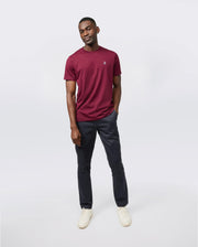 A person is standing against a plain white background. They are wearing a maroon short-sleeve men's crewneck tee, dark blue trousers, and white sneakers. The Psycho Bunny MENS CLASSIC CREW NECK TEE - B6U014Z1PC looks breathable and lightweight. With one hand in their pocket and a smile, they exude a casual and relaxed vibe.