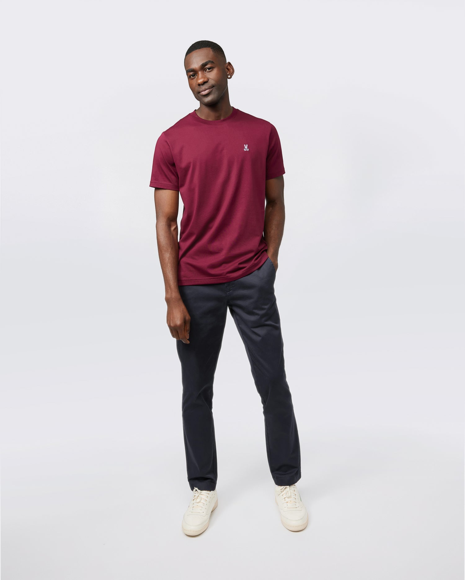A person is standing against a plain white background. They are wearing a maroon short-sleeve men's crewneck tee, dark blue trousers, and white sneakers. The Psycho Bunny MENS CLASSIC CREW NECK TEE - B6U014Z1PC looks breathable and lightweight. With one hand in their pocket and a smile, they exude a casual and relaxed vibe.