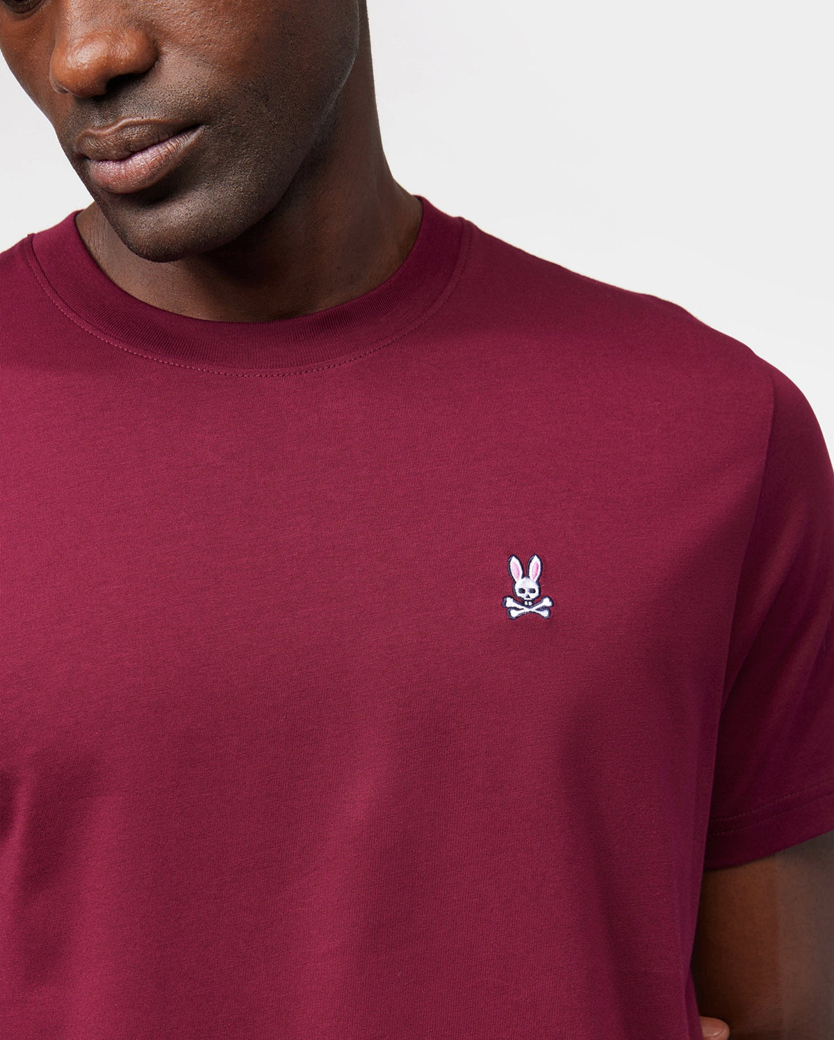 A person wearing a burgundy Psycho Bunny MENS CLASSIC CREW NECK TEE - B6U014Z1PC made from breathable and lightweight Pima cotton with a small white embroidered bunny and crossbones logo on the left side of the chest. The background is plain white, and the person's face is partially visible.