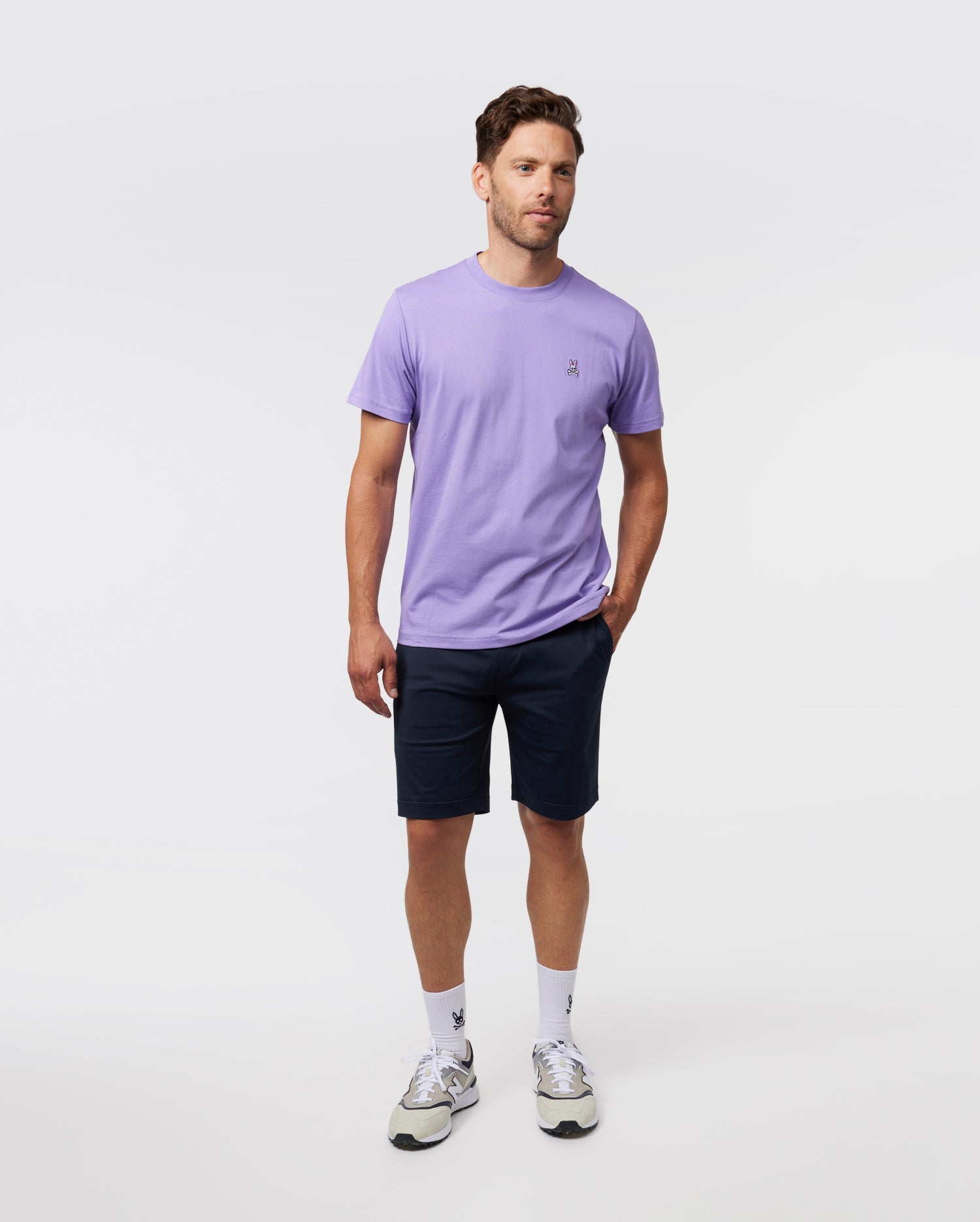 A man in a lavender Psycho Bunny MENS CLASSIC CREW NECK TEE - B6U014Z1PC and navy shorts stands against a plain white background. He's wearing white socks and gray sneakers, with a relaxed posture.