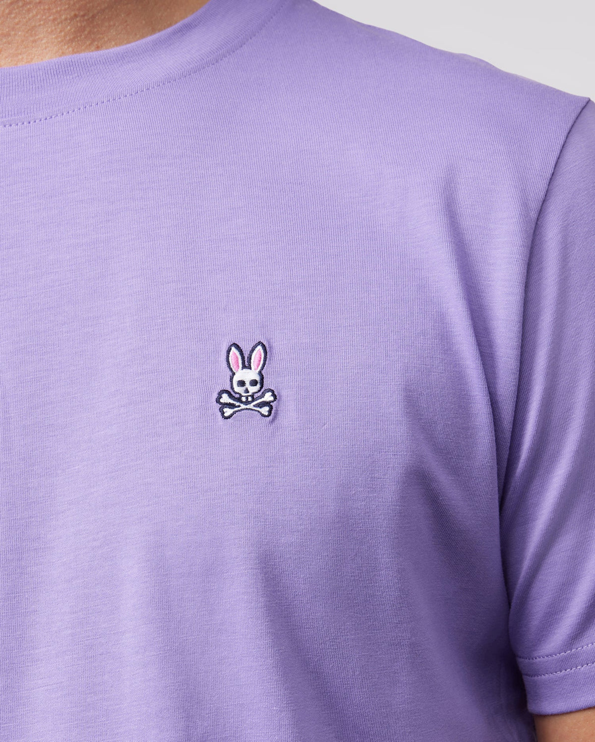 A close-up showcases an individual donning the Psycho Bunny Men's Classic Crew Neck Tee - B6U014Z1PC in light purple, crafted from soft Pima cotton. The left chest area features a distinctive small embroidered bunny logo with pink ears above crossed bones. A neutral background emphasizes this unique shirt design.