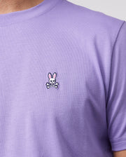 A close-up showcases an individual donning the Psycho Bunny Men's Classic Crew Neck Tee - B6U014Z1PC in light purple, crafted from soft Pima cotton. The left chest area features a distinctive small embroidered bunny logo with pink ears above crossed bones. A neutral background emphasizes this unique shirt design.