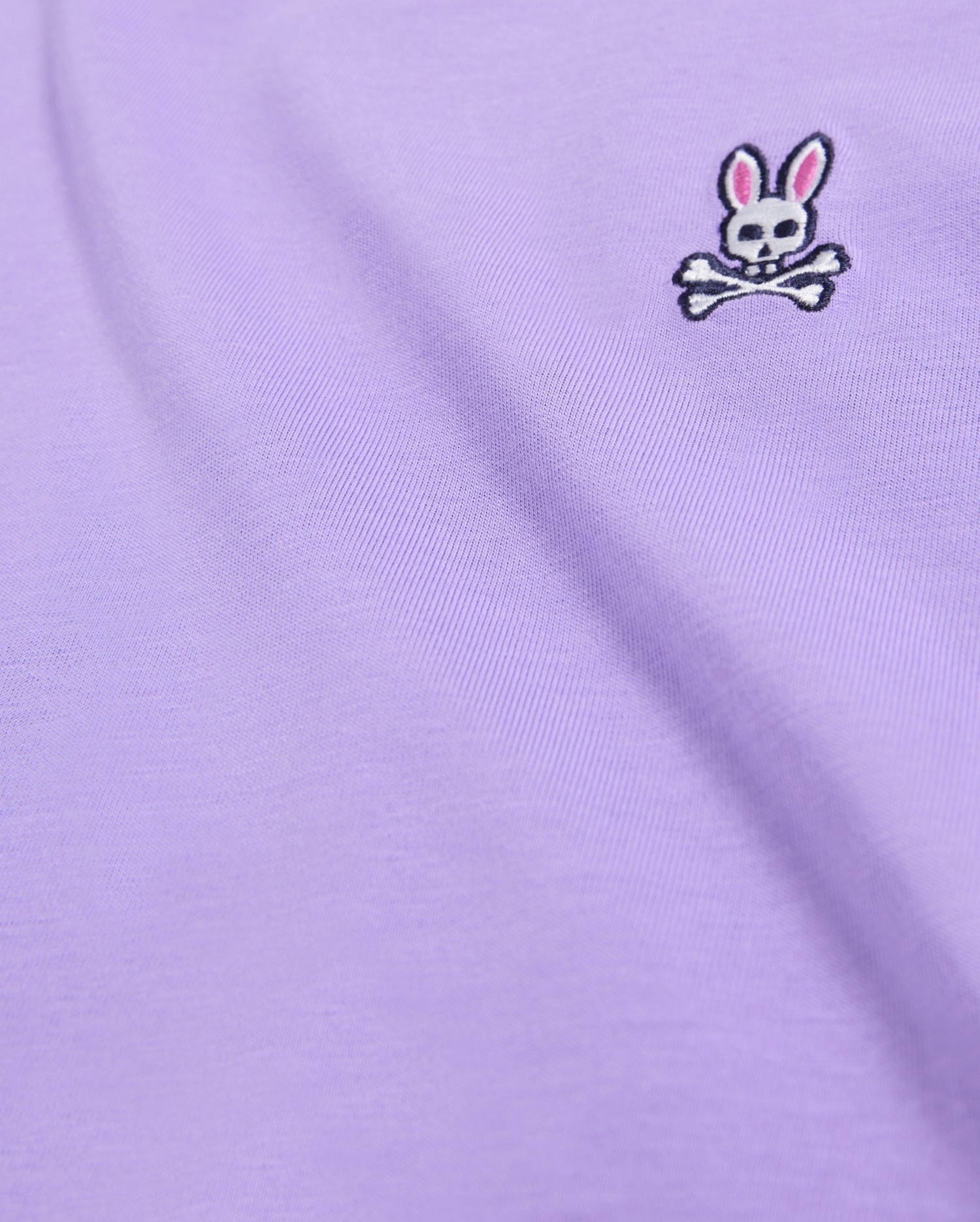 A close-up of a lavender-colored fabric, likely from the Psycho Bunny Men's Classic Crew Neck Tee (B6U014Z1PC), showcases a small embroidered bunny logo with skull and crossbones. The rabbit sports pink ears and black eyes on a smooth, slightly draped Pima cotton material.