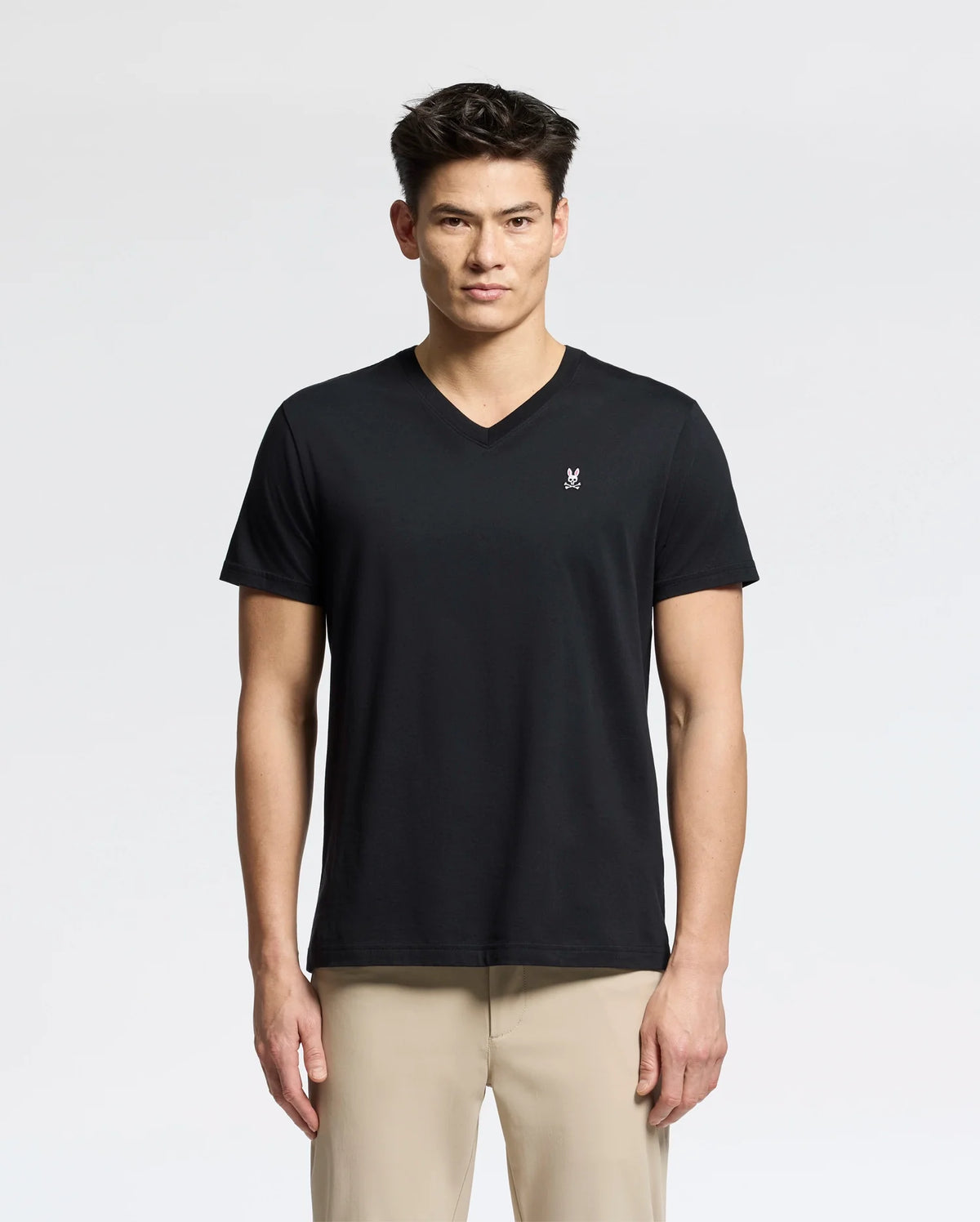 A person in the Psycho Bunny MENS CLASSIC V NECK TEE - B6U100ARPC, made of soft Pima cotton and featuring a small white chest logo, pairs it with light beige pants against a plain white background. They have a neutral expression while wearing this black tee.