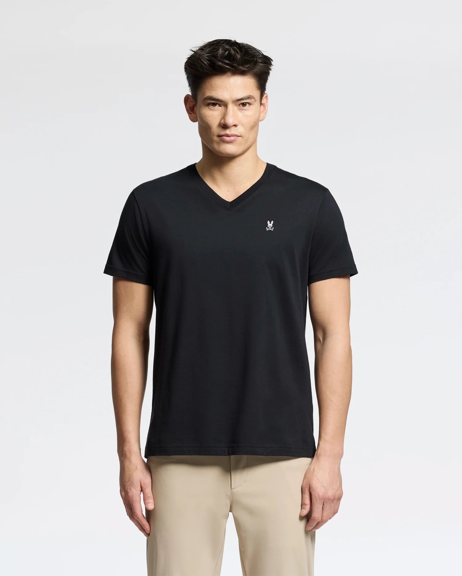 A person in the Psycho Bunny MENS CLASSIC V NECK TEE - B6U100ARPC, made of soft Pima cotton and featuring a small white chest logo, pairs it with light beige pants against a plain white background. They have a neutral expression while wearing this black tee.