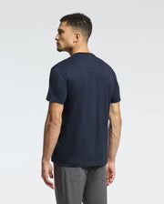 A man stands with his back to the camera, wearing a Psycho Bunny MENS CLASSIC V NECK TEE - B6U100ARPC in navy and gray pants. The background is a simple, light gray.