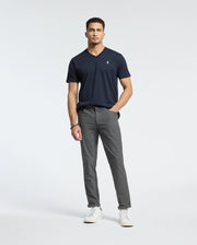A man stands against a plain background, wearing the Psycho Bunny MENS CLASSIC V NECK TEE (B6U100ARPC) in dark blue, crafted from soft Pima cotton. He pairs it with gray pants and white sneakers. With short curly hair and a relaxed posture, he looks directly at the camera.