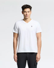 Against a light background, a person wears the Psycho Bunny Men's Classic V-Neck Tee in white with an embroidered logo, paired with black pants. The 100% Pima cotton tee complements their short dark hair as they look directly at the camera with a neutral expression.