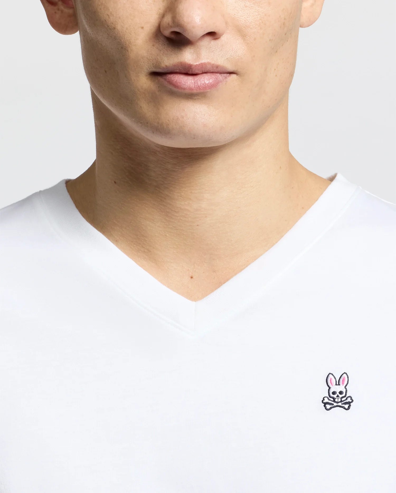 Someone wears a Men's Classic V Neck Tee - B6U100ARPC by Psycho Bunny, made of 100% Pima cotton, displaying the Psycho Bunny logo with an embroidered pink bunny and crossed bones on the left chest against a plain white background.