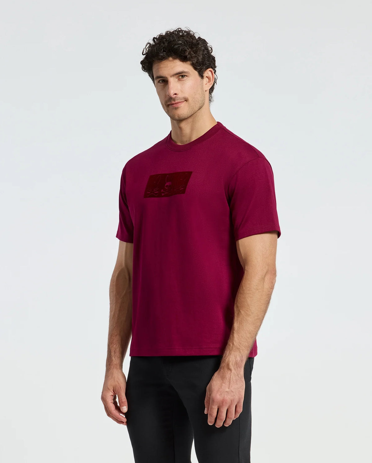 A man wearing a Psycho Bunny MENS ROMEO HEAVY WEIGHT RELAXED FIT TEE in maroon stands against a plain background. He has curly dark hair and pairs the shirt with black pants. With its comfortable relaxed fit, he looks directly at the camera in a casual posture.