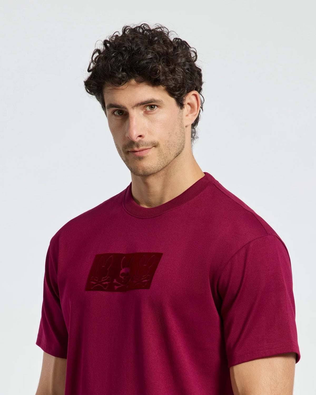 A person with short curly hair is wearing a maroon Psycho Bunny MENS ROMEO HEAVY WEIGHT RELAXED FIT TEE - B6U110E200, featuring a rectangular design on the chest. They are standing against a plain light background, looking at the camera with a neutral expression.
