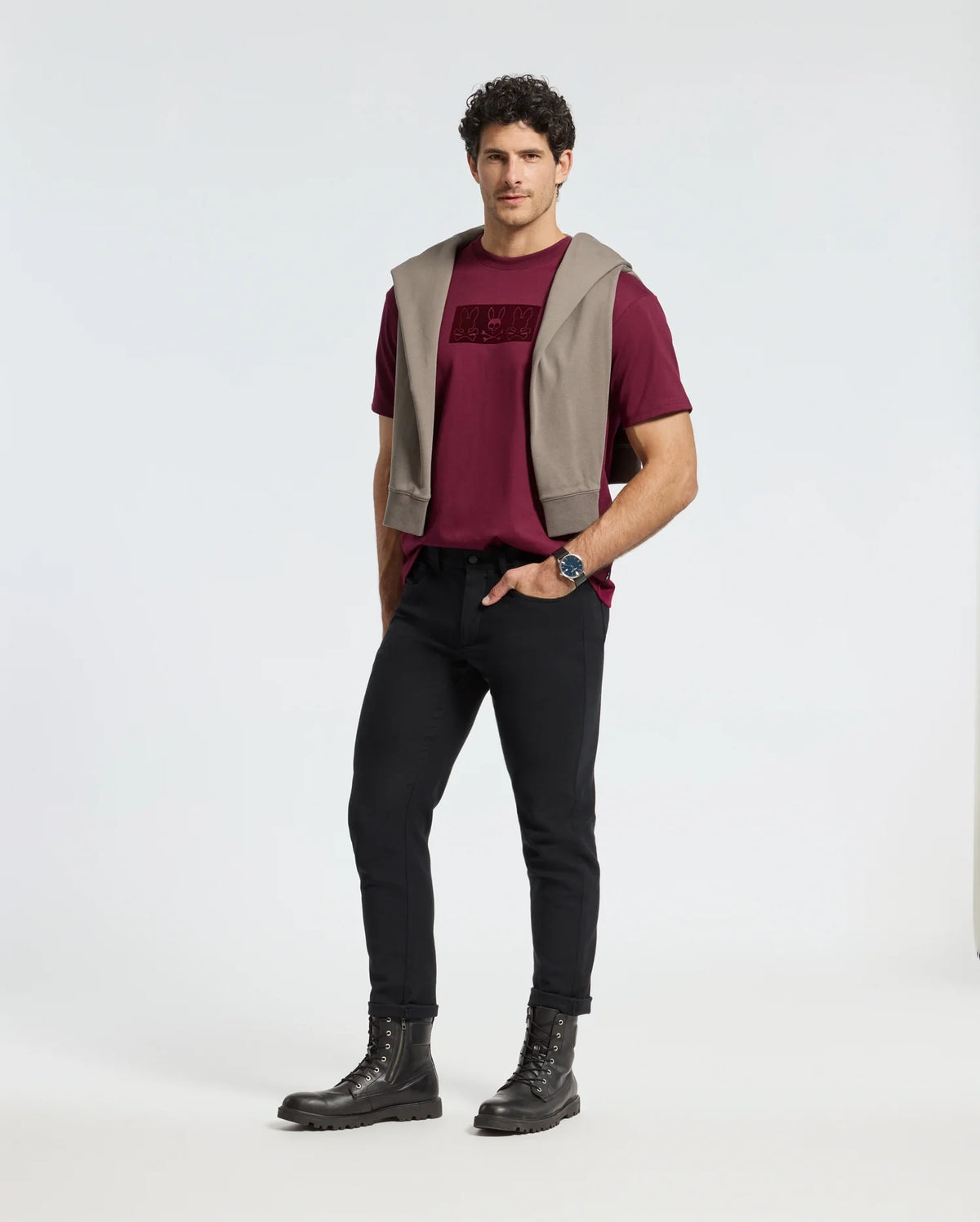 A man in a burgundy Psycho Bunny MENS ROMEO HEAVY WEIGHT RELAXED FIT TEE - B6U110E200 and black pants, with a gray sweater draped over his shoulders, stands against a plain background. With hands in pockets, he wears comfortable black boots and a watch, exuding an effortlessly relaxed fit.