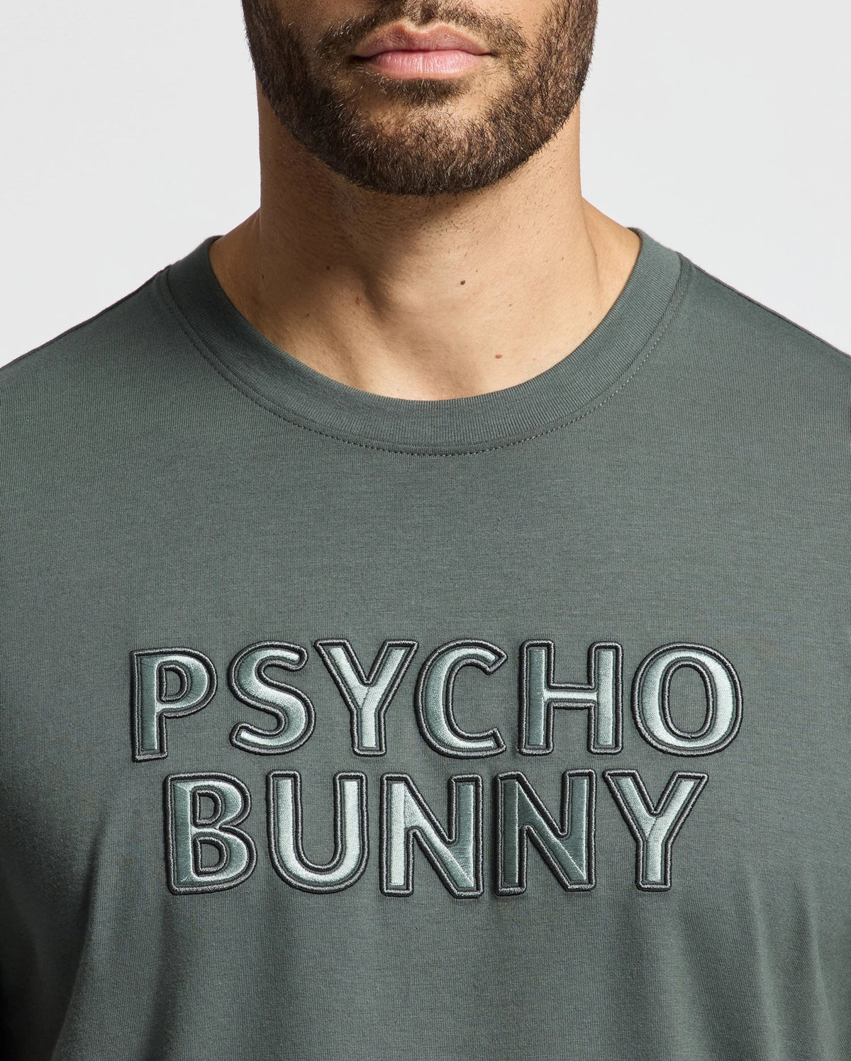 A person wearing a MENS PIERCE EMBROIDERED GRAPHIC TEE by Psycho Bunny featuring an eye-catching green color and a striking "Psycho Bunny" graphic boldly embroidered across the chest. The vibrant hue of this tee stands out against the white background.