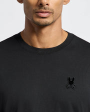 A person wearing the Psycho Bunny Men's Branson Fashion Tee in plain black, featuring a small, stylized rabbit skull and crossbones logo on the upper left side. This premium quality, lightweight T-shirt stands out against a plain and neutral background.