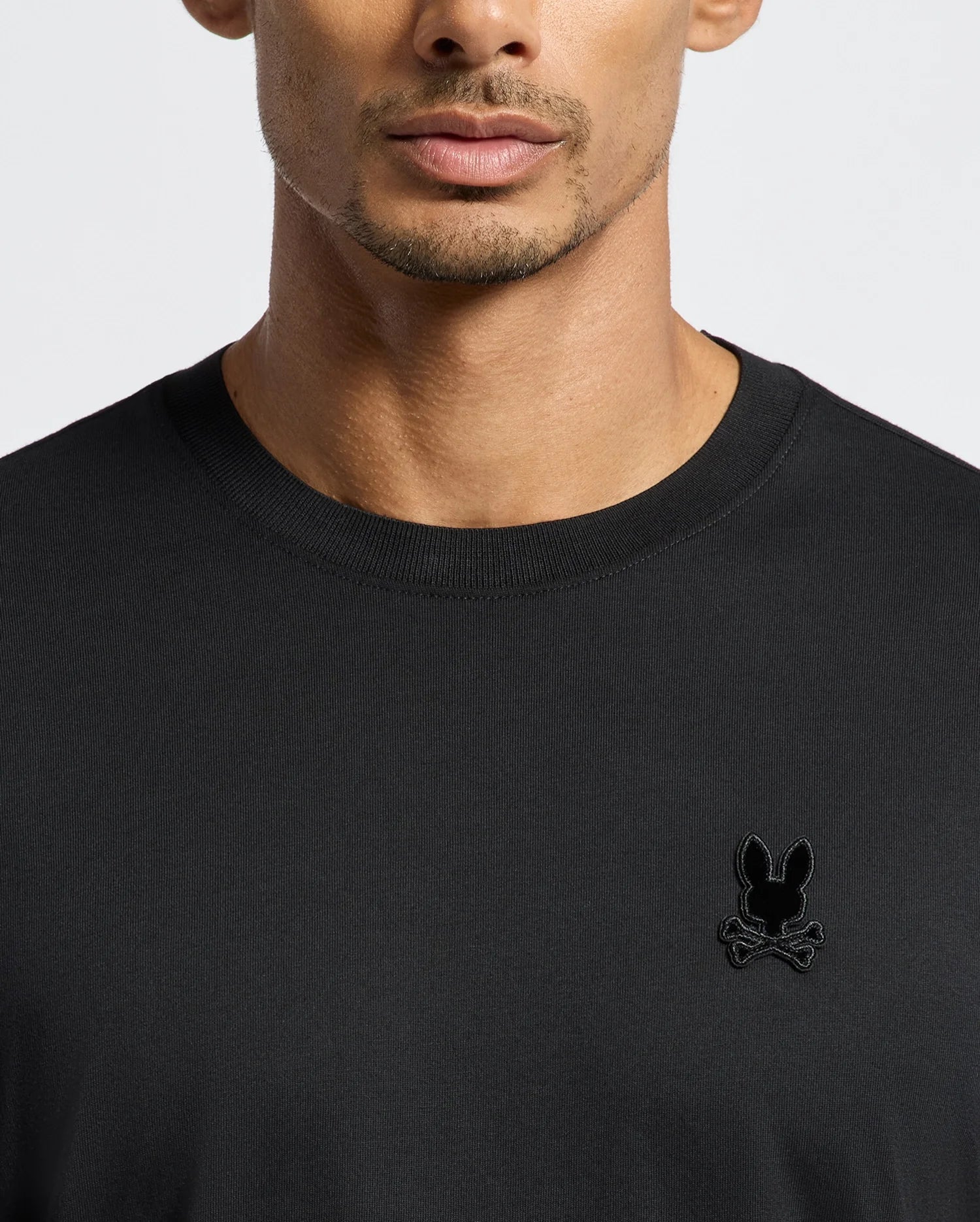 A person wearing the Psycho Bunny Men's Branson Fashion Tee in plain black, featuring a small, stylized rabbit skull and crossbones logo on the upper left side. This premium quality, lightweight T-shirt stands out against a plain and neutral background.