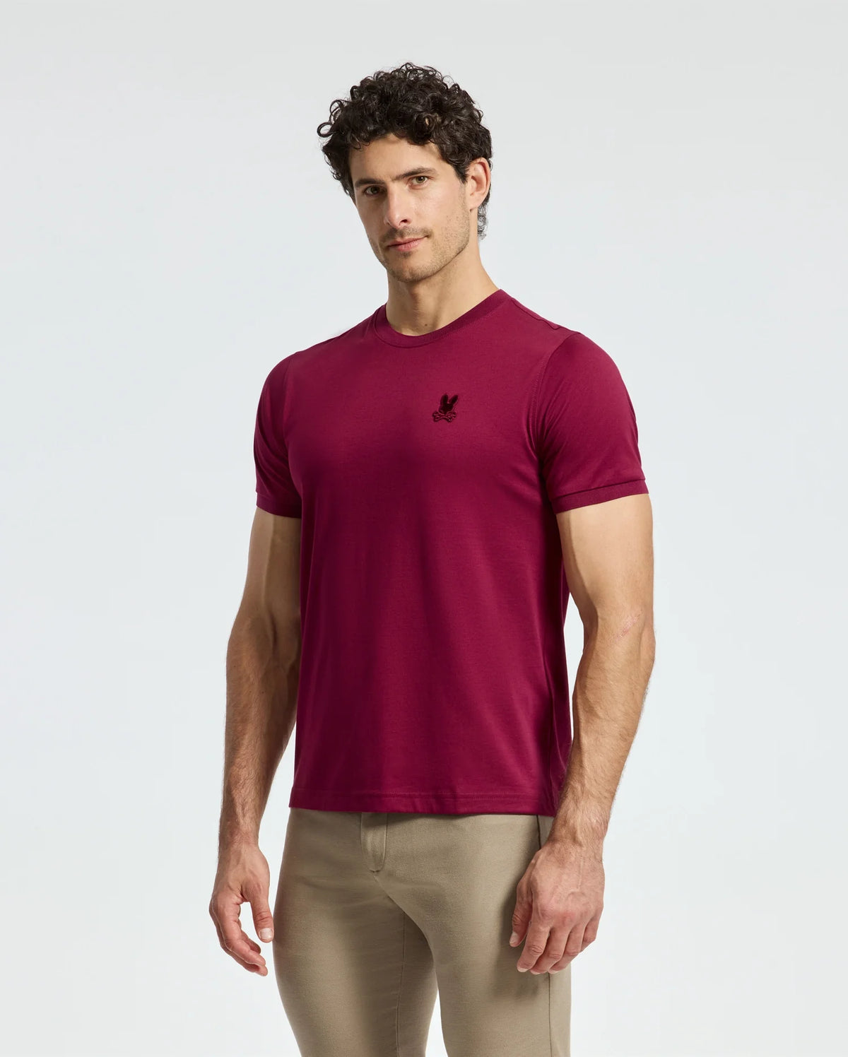 A man wearing a tailored maroon Psycho Bunny Men's Branson Fashion Tee - B6U137E200, featuring a small logo on the chest, stands against a plain background. He also has on beige pants, with his hands relaxed by his sides.