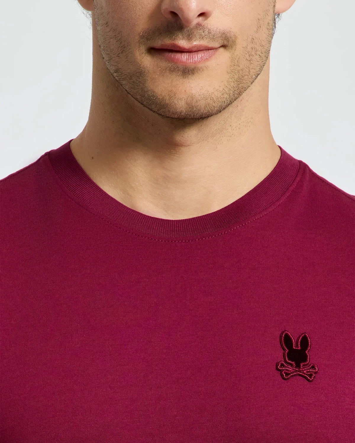 A person is seen in a stylish Psycho Bunny MENS BRANSON FASHION TEE - B6U137E200, crafted from lightweight cotton in a vibrant magenta color. The tee features a distinctive black embroidered bunny logo on the chest. The lower half of the face and upper torso are visible against a plain background, highlighting the tee's chic and effortless style.