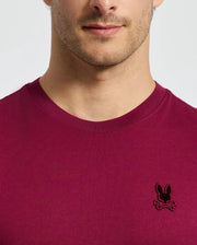 A person is seen in a stylish Psycho Bunny MENS BRANSON FASHION TEE - B6U137E200, crafted from lightweight cotton in a vibrant magenta color. The tee features a distinctive black embroidered bunny logo on the chest. The lower half of the face and upper torso are visible against a plain background, highlighting the tee's chic and effortless style.
