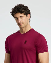 A man with curly hair is wearing a deep red Psycho Bunny MENS BRANSON FASHION TEE - B6U137E200, featuring a small embroidered logo on the chest. He stands against a plain background, looking directly at the camera.