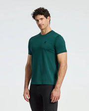 A man with curly hair is wearing a green Psycho Bunny MENS BRANSON FASHION TEE - B6U137E200 and black pants, standing against a plain white background. The tee, renowned for its lightweight comfort, features a small logo on the chest.