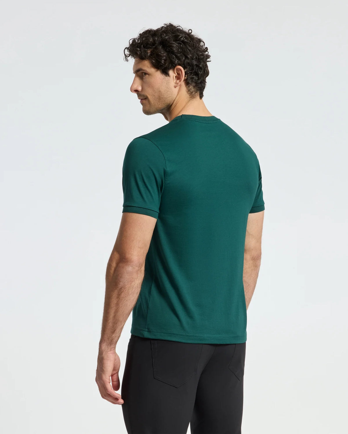 A man with curly hair stands facing away from the camera, wearing a green short-sleeved Psycho Bunny Men's Branson Fashion Tee - B6U137E200 and black pants against a plain white background.