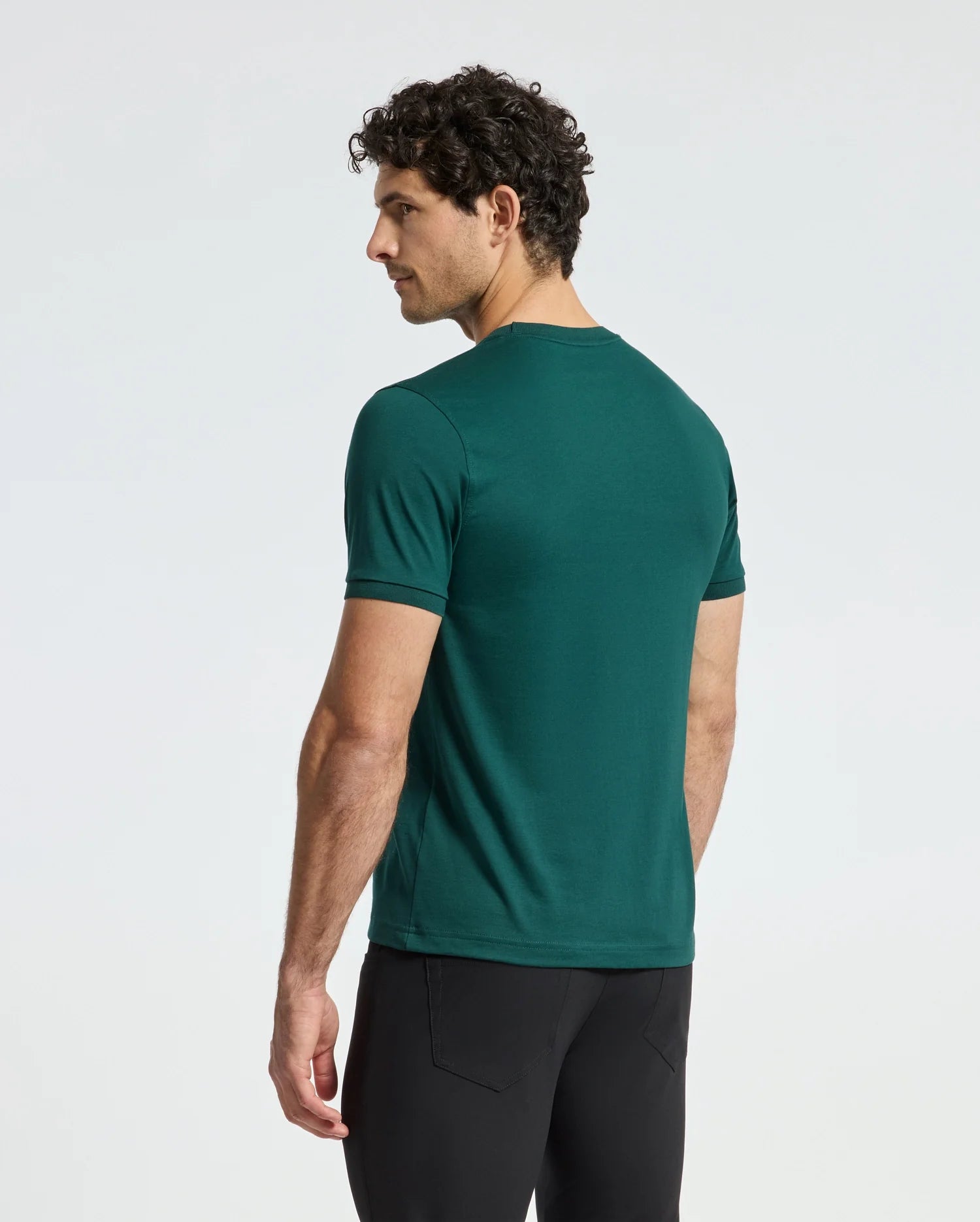 A man with curly hair stands facing away from the camera, wearing a green short-sleeved Psycho Bunny Men's Branson Fashion Tee - B6U137E200 and black pants against a plain white background.
