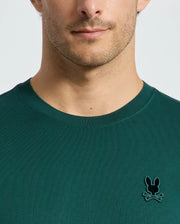 A person wearing a dark green Psycho Bunny MENS BRANSON FASHION TEE - B6U137E200, featuring a small embroidered emblem of a bunny and crossbones on the chest. The focus is on the shirt's lightweight fabric and design, rather than the person's face.