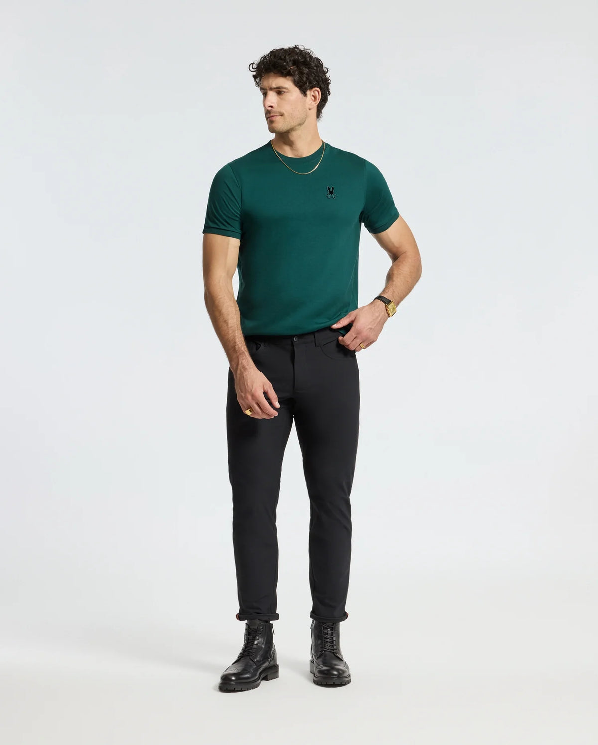 A man wearing a Men's Branson Fashion Tee from Psycho Bunny, paired with black pants and black boots, stands against a plain white background. He is looking to the side with a casual posture and is accessorized with a gold necklace and bracelet.