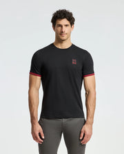 A man with curly hair is wearing the Psycho Bunny MENS PIERCE FASHION TEE (B6U140E200), a black Pima cotton tee featuring red trim on the sleeves and an embroidered Bunny patch on the chest. He stands facing forward with his hands at his sides against a plain light background.