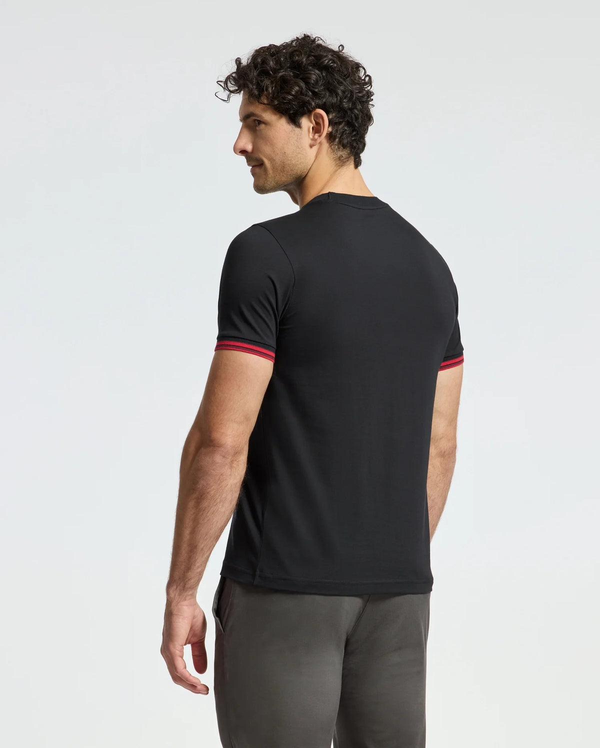 A man with curly hair slightly turns to the side, wearing a black MENS PIERCE FASHION TEE - B6U140E200 by Psycho Bunny, which features red-trimmed sleeves and an embroidered Bunny patch. He pairs it with dark pants and stands against a plain white background.