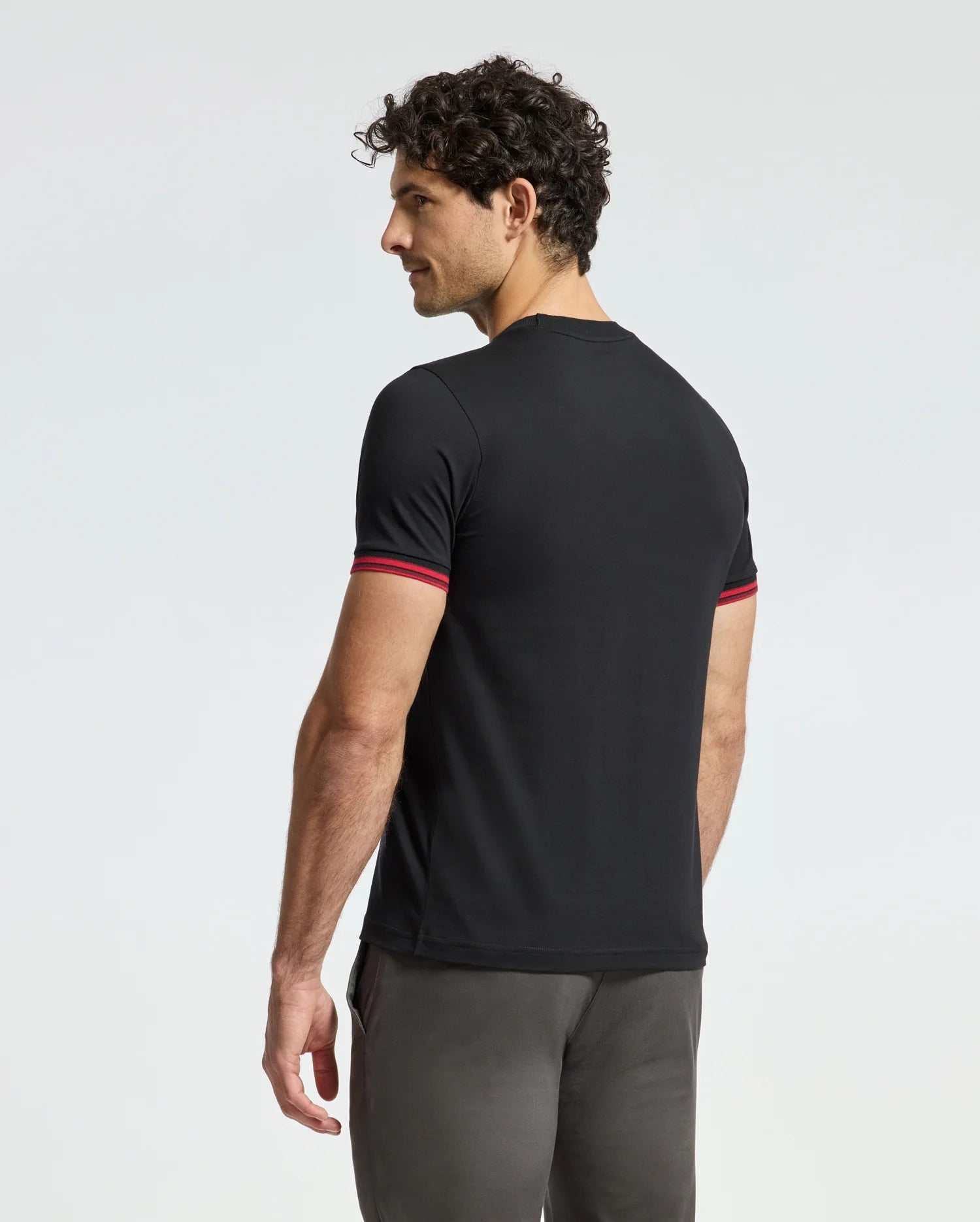 A man with curly hair slightly turns to the side, wearing a black MENS PIERCE FASHION TEE - B6U140E200 by Psycho Bunny, which features red-trimmed sleeves and an embroidered Bunny patch. He pairs it with dark pants and stands against a plain white background.