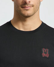 A model wears the Psycho Bunny Men's Pierce Fashion Tee (B6U140E200), made from soft Pima cotton and highlighting an embroidered Bunny patch with crossed bones on the chest. This black T-shirt features a design that is both subtle and stylish, displaying only the lower half of the face and upper torso.