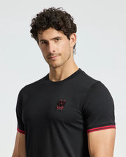 A man with curly hair wears the fitted black MENS PIERCE FASHION TEE - B6U140E200 from Psycho Bunny, featuring red trim on the sleeves and a small embroidered Bunny patch on the chest. He stands against a plain light background.